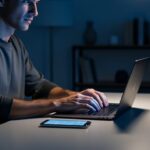 A person typing on a laptop in a dim home office at night, with a softly lit smartphone nearby to suggest online privacy needs.